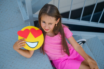 A winking Girl sitting on the stairs shows emojis with loving eyes and big hearts. World Smiley Day