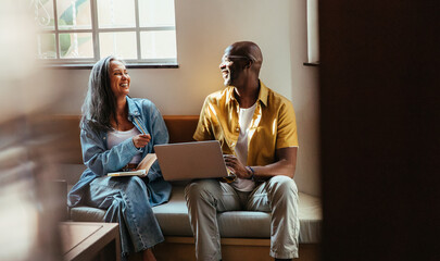 Happy colleagues laughing and collaborating while sitting on a couch with a laptop in a modern...