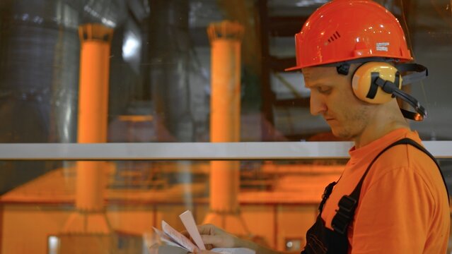 Factory worker cross-checks data with checklist during night shift. Ensuring accuracy and quality control in production. Dressed in safety equipment, focused on maintaining standards.