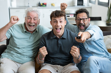 Enthusiastic three generation men family fans watch football championship or olympic games on TV sitting together on sofa, happy grandson with father and old grandfather celebrate team victory