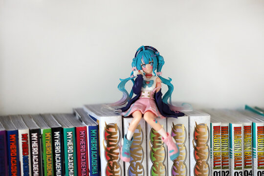 
Hatsune Miku figure sitting in a bookcase on manga books. Virtual singer with vocaloid. Japanese manga and anime style doll.