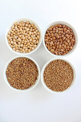 Four white bowls filled with chickpeas, kidney beans, lentils and buckwheat arranged on a white background
