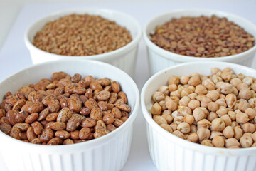 Four white bowls are filled with various legumes including chickpeas, lentils, beans, and buckwheat groats