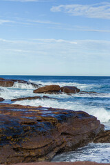 The image captures a serene coastal scene with rocky formations by the sea, gentle waves lapping against the shore, and a clear blue sky with scattered clouds in the background.