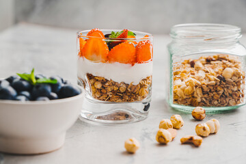 Healthy breakfast, parfait dessert with granola, Greek yogurt and strawberries and blueberries in a transparent glass on a gray background with ingredients. Selected focus