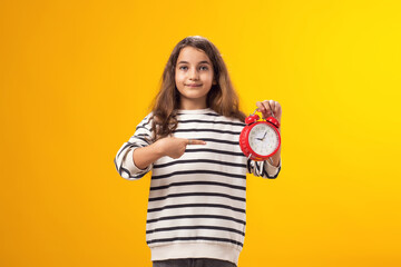 Smiling kid girl holding an alarm clock in hand and pointing finger at it. The concept of education, school, deadlines, time to study