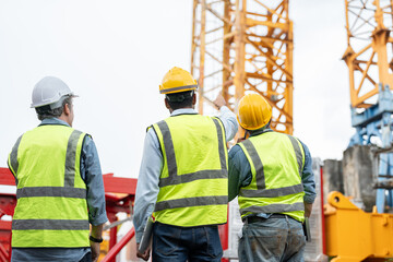 Group of male engineer wearing safety vests and helmet pointing to tower crane for surveying and...