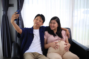 Happy man with pregnant wife rubbing his belly while relaxing and spending time in modern apartment. Pregnant woman and husband spending time together using smartphone