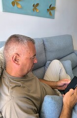 Fototapeta premium Young handsome adult man sitting on sofa chatting with smartphone.Boy sitting on sofa with mobile phone.Happy man on sofa looking at mobile phone.Relaxed boy with mobile phone on sofa