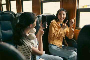 Two female friends are sitting in a train, one is pointing at the window and the other is taking a picture with her phone