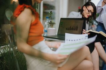 Focused businesswomen collaborating in an outdoor meeting, analyzing data on a laptop and printed graphs. Professional teamwork and modern work environment concept.