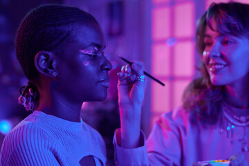 Fototapeta premium Side view portrait of Black young woman trying luminescent make up look in dark getting ready for party, copy space