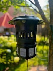 A bug Zapper hangs from a tree outside, and attracts bugs, insects and mosquitoes using a purple...