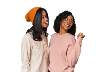 Young Latin friends together in studio points with thumb finger away, laughing and carefree.