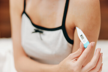 Woman holding electronic thermometer sitting on bed in room with white bedspread