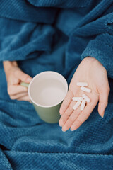 Person holding cup of pills and coffee wrapped in blanket with pillows