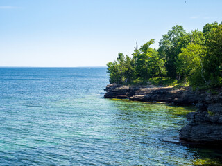 Cliffs and green vegetation by the clear water of Lake Ontario, New York state, with blue sky and horizon line.