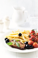 Roast Pork Steak with french fries, tomatoes and olives. White wooden background. Close up.	