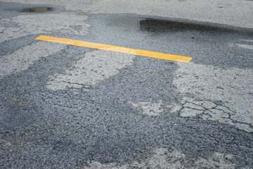 Fototapeta premium A yellow line is painted on a wet road. The road is cracked and has potholes