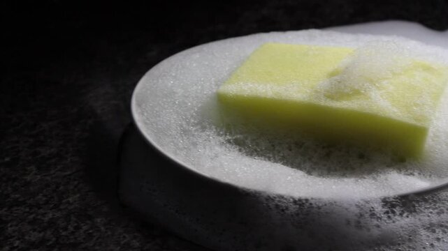 Un primer plano de una esponja amarilla llena de jab&oacute;n liquido en un plato de color blanco en la cocina.