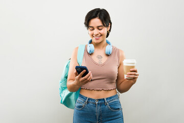 Joyful young woman with headphones, backpack, smartphone, and coffee in a studio