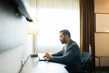 Profile view of a young entrepreneur is seated at a desk in his hotel room, using a laptop