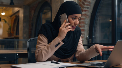 Muslim woman in hijab female business employer consultant office manager Arabian smiling businesswoman talk mobile phone consult at workplace happy positive islamic girl looking at laptop online order
