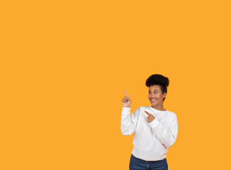 A young woman with short curly hair is smiling and pointing at copy space while standing in front of a solid yellow background.