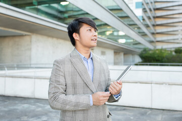 Fototapeta premium Asian businessman stands in front of a modern building, holding a tablet in his hands. He is looking up, seemingly in deep thought or listening to someone off-screen, copy space