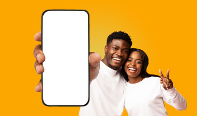 A young black couple smiles brightly as they hold a smartphone with a blank white screen in front of them. The woman makes a peace sign with her hand, while the man points at the screen.
