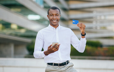 African American young man in a white shirt and grey pants stands outside with a blue credit card in one hand and a smartphone in the other. He smiles brightly as he looks directly at the camera.