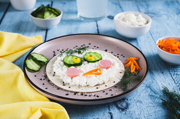 Face made of tortilla, cottage cheese, sausages, vegetables for children's breakfast on a plate