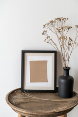 A black picture frame with blank white paper inside is sitting on top of an old wooden table, and there are dried flowers in the vase next to it. The background wall has no decorations or shadows.