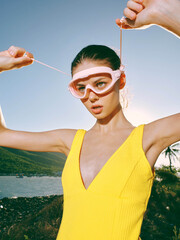 A woman in a yellow dress holds a pair of goggles on her head, looking joyful and playful, against...