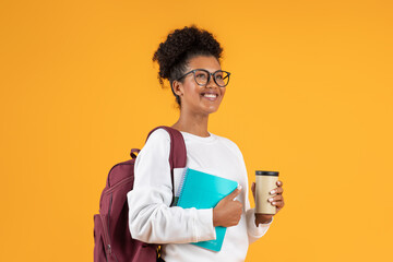 A young woman with a backpack, glasses, and a bright smile holds a blue notebook and a cup of coffee while standing in front of a bright yellow backdrop.