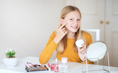 A teen girl with long, blonde hair sits at a vanity table, applying a facial cream to her cheek....