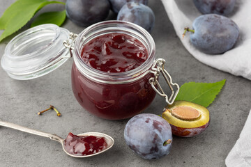Sweet plum jam or marmalade in glass jar  closeup and fresh raw whole and sliced fruits with leaves on dark grey stone table background