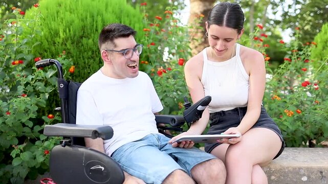 A young girl cares for an adult man with cerebral palsy in a park at sunset.The young woman shows the disabled man a tablet to entertain him.Concept of people with dependence due to cerebral palsy.