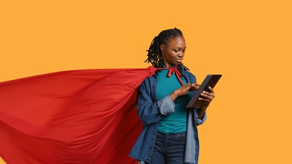 Joyous girl wearing superhero costume excited to purchase stuff online using tablet, studio background. Teenager dressed as comic book hero happy to use credit card for internet shopping, camera B © DC Studio