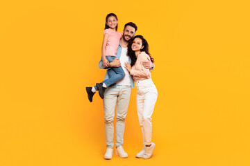 A smiling family stands together against a bright yellow background. The father holds his daughter in his arms, while the mother stands beside him, her arm around his waist