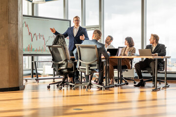 A businessman in a suit stands at the front of a conference table, presenting financial data on a large screen to a team of colleagues