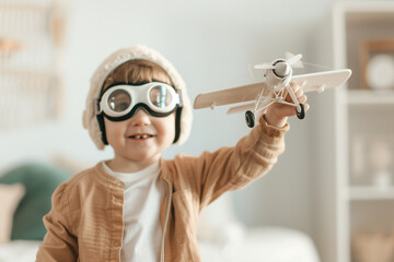 Joyful toddler boy dressed as an aviator pilot, wearing goggles and holding a toy airplane in his room. Child's imagination, dream, playful freedom, creative playtime photo