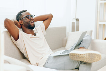 Happy African American Freelancer Working on Laptop while Sitting on Sofa in Modern Living Room He is Typing and Smiling, Enjoying the Comfort of His Home The Cyberspace and Technology Enhance His