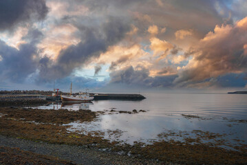 A tranquil harbor in Iceland with a docked fishing boat, colorful clouds reflecting on calm waters, and a shoreline covered in seaweed and rocks.