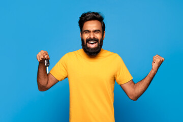 Fototapeta premium Hindu man with a beard is smiling and holding up a car key in his right hand while his left fist is clenched in the air. He is wearing a yellow t-shirt and is standing in front of a blue background.