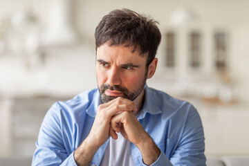 Naklejka premium A man with short brown hair and a beard is sitting indoors, wearing a blue button-down shirt. He is looking off to the side, resting his chin on his hands, and appears to be deep in thought.