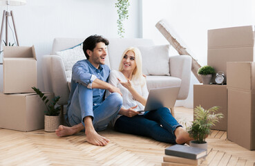A couple sits on the floor of their new home, surrounded by unpacked boxes, while planning on a laptop.