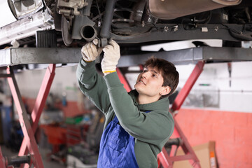 Young guy mechanic in uniform repairs underbody of car in car service station