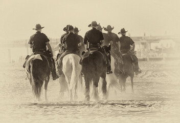 A Group of Police Officers on horseback on beach in late afternoon sun. The image is in black and...