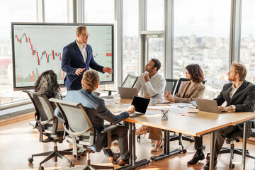 A businessman is presenting financial data to a team in a modern office with large windows overlooking the city. He is standing and pointing at a chart on a screen